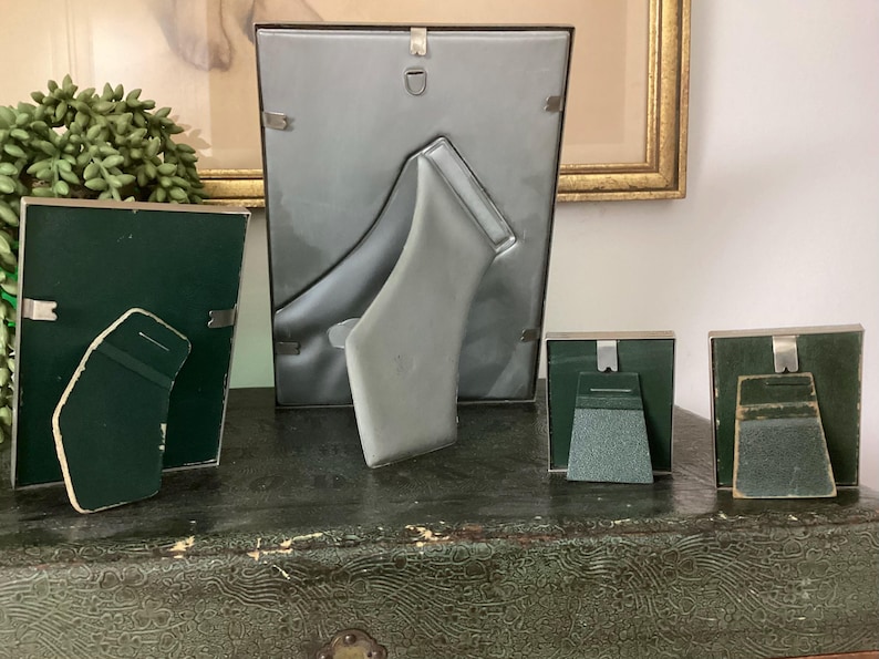 May include: Four silver picture frames with green felt backing. The frames are arranged in a row on a green surface. The largest frame is standing upright, while the three smaller frames are lying flat.