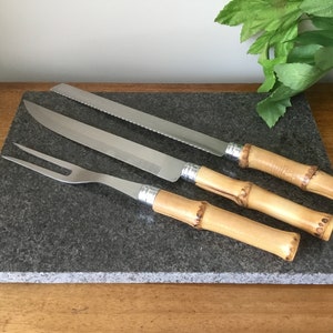 Op de afbeelding: Drie keukmessen met bamboe handvatten op een grijs stenen snijplank. De messen hebben zilveren bladen en zijn in een rij gerangschikt.