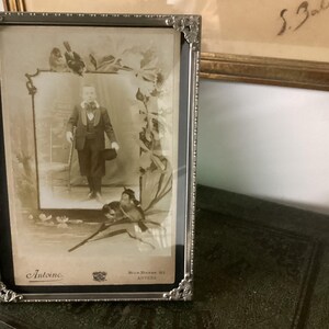 May include: A silver-toned picture frame with a black mat and a sepia-toned photograph of a young boy in a suit and hat. The photo is surrounded by a decorative border of birds and leaves. The frame has a label that reads "Antoine" and "Rue Notre Dame 33, Anvers".
