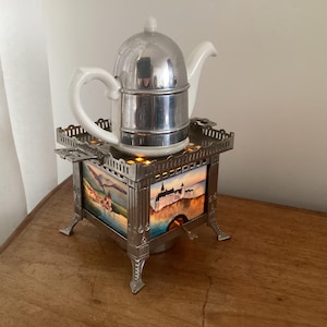 May include: A vintage silver tea kettle with a white handle and spout, on a decorative metal stand. The stand has a painted landscape scene and ornate detailing. A unique tea set, perfect for a kitchen display.