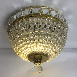May include: A gold-toned ceiling light fixture with a round, crystal-covered shade. The shade is made up of many small, clear crystals that are arranged in a circular pattern. The light fixture has a single bulb in the center.