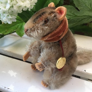May include: A brown and gray plush squirrel wearing a red velvet scarf with a gold button. The squirrel is sitting on a white surface.