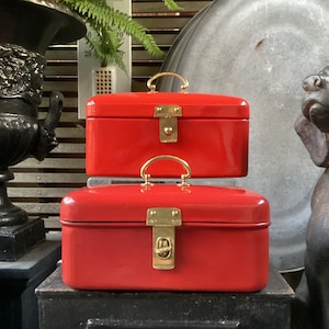 May include: Two red rectangular boxes stacked on top of each other. Each box has a gold-colored handle and a gold-colored clasp. The boxes are sitting on a black surface, with a decorative urn and a dog statue in the background.