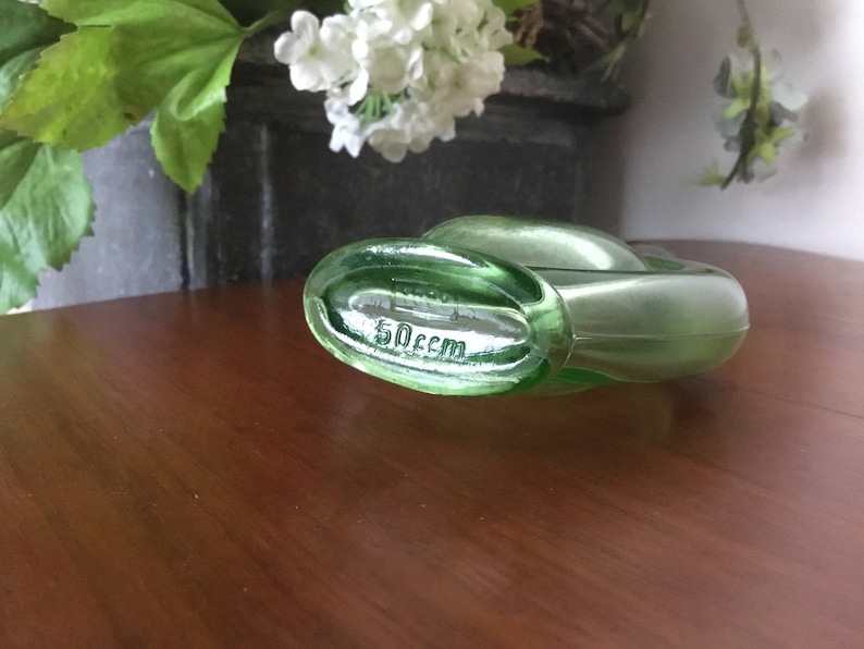 May include: A green glass bottle with a unique shape and a label that reads "50ccm".