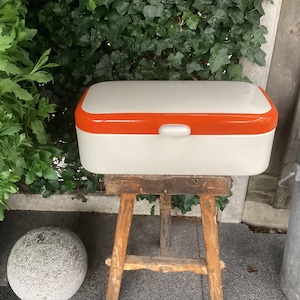 Può includere: Una scatola per il pane bianca con un coperchio arancione. La scatola per il pane è appoggiata su uno sgabello di legno.