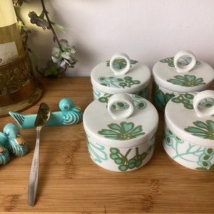 以下が含まれることがあります： 緑と青の花柄が描かれた白いセラミック製の容器4つ。白い蓋にはリング状の取っ手が付いています。容器は木製の表面に正方形に並べてあります。