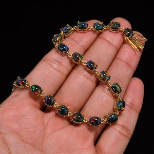 May include: A gold-toned link bracelet featuring a series of oval-shaped black opals. Each opal displays iridescent flashes of red, green, and blue color. The bracelet is secured with a gold-toned metal clasp mechanism at one end of the chain.