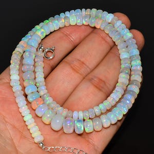 Puede incluir: Collar compuesto por cuentas de ópalo iridiscentes de varias formas y tamaños. Las cuentas están ensartadas con un cierre y una cadena de plata. El collar se muestra en una mano sobre un fondo negro.