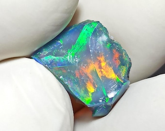 Puede incluir: Una gema de ópalo en bruto con colores vibrantes de verde, naranja y azul. La gema se sostiene entre dos manos enguantadas de blanco, mostrando su forma natural e irregular y su juego de colores iridiscentes. El fondo es negro.