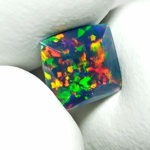 May include: A square-cut black opal gemstone featuring a dark base color with vibrant, multi-colored flashes of red, orange, yellow, and green throughout its surface. The gemstone has a faceted top and is held between two white textured surfaces.