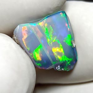 Könnte beinhalten: Ein polierter Opal-Edelstein mit einer lebendigen Farbgebung. Der Stein zeigt grüne, gelbe und blaue Lichtblitze vor einem dunklen Hintergrund. Der Opal wird von einer weißen Oberfläche gehalten.