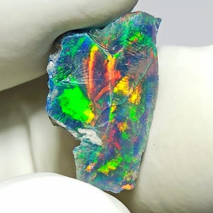 May include: A raw, uncut opal gemstone featuring a dark, translucent base with vibrant flashes of neon green, bright yellow, and fiery orange colour play across its irregular, fractured surface, showcasing natural geological textures and patterns.