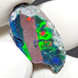 Puede incluir: Una gema de ópalo en bruto con una vibrante exhibición de color. La piedra presenta destellos de verde, rojo y azul sobre un fondo oscuro. El ópalo se sostiene en una mano enguantada, mostrando su forma natural e irregular.