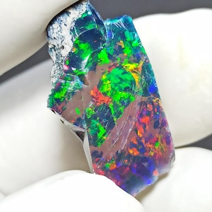 Puede incluir: Una vibrante gema de ópalo en bruto con un color base oscuro, que muestra un juego de colores con destellos de verde, rojo y amarillo. La piedra en bruto es sostenida por una mano enguantada, mostrando su textura natural, sin pulir y sus cualidades iridiscentes.