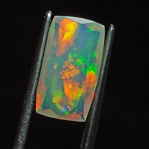 May include: A rectangular, iridescent opal gemstone with flashes of green, orange, and red. The opal is held in tweezers against a black background.