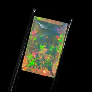 Puede incluir: Una piedra preciosa de ópalo rectangular de color claro con destellos verdes, rojos y naranjas. La piedra preciosa está sujeta con unas pinzas.