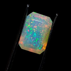 Puede incluir: Una piedra preciosa de ópalo blanco con destellos verdes y naranjas. La piedra preciosa está tallada en forma rectangular y se sostiene con unas pinzas.