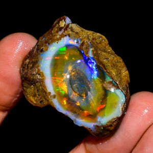 Könnte beinhalten: Ein roher Opal-Edelstein mit einer lebendigen Farbgebung. Der Stein weist eine Mischung aus gelben, grünen, blauen und orangen Farbtönen mit einer weißen und braunen Außenschicht auf. Der Opal wird in einer Hand vor schwarzem Hintergrund gehalten.
