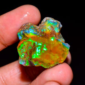 May include: A raw, unpolished opal gemstone displaying a vibrant play-of-color with intense flashes of neon green, bright orange, and deep blue. The stone has an irregular, organic shape with a translucent, glassy texture and visible inclusions.