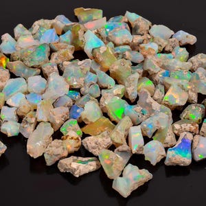 May include: A collection of raw opal gemstones. The opals display a variety of colors, including iridescent flashes of blue, green, yellow, and orange. The rough stones have a natural, unpolished appearance, with a mix of white, tan, and gray hues.