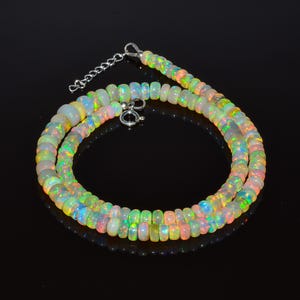 May include: A necklace featuring iridescent opal beads. The beads display a mix of white, yellow, green, and pink, with flashes of blue and orange. The necklace is secured with a silver clasp and chain.
