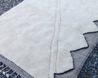 Könnte beinhalten: Ein cremefarbener, rechteckiger Teppich mit geometrischem Design. Der Teppich hat einen dicken, strukturierten Flor und einen schwarz-weißen Zickzack-Rand. Quasten schmücken den Rand. Der Teppich ist für ein Wohn- oder Schlafzimmer geeignet.