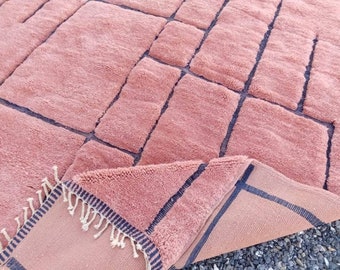 Puede incluir: Una alfombra grande y rectangular de color rosa con un patrón geométrico de líneas azul oscuro. La alfombra tiene un pelo grueso y texturizado y un borde con flecos. La alfombra está doblada, revelando la parte inferior.