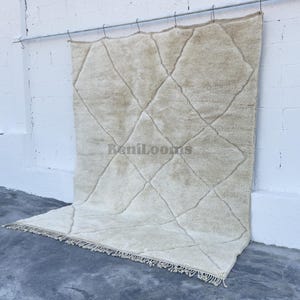 May include: A large, rectangular, off-white area rug featuring a repeating diamond pattern formed by thin, dark lines. The rug has a thick, textured pile and a fringe along the bottom edge. The text "BeniLooms" is printed in the center of the rug.