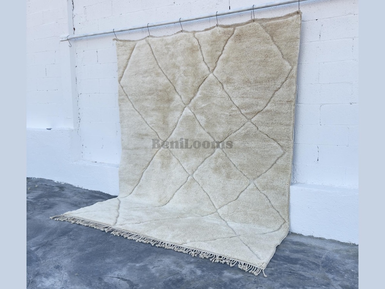 May include: A large, rectangular, off-white area rug featuring a repeating diamond pattern formed by thin, dark lines. The rug has a thick, textured pile and a fringe along the bottom edge. The text "BeniLooms" is printed in the center of the rug.