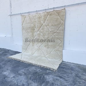 May include: A large, rectangular, cream-colored wool rug featuring a repeating diamond pattern formed by thin, dark lines. The rug has a thick, shaggy pile texture and includes a fringe along the bottom edge. The text BeniLooms is visible.