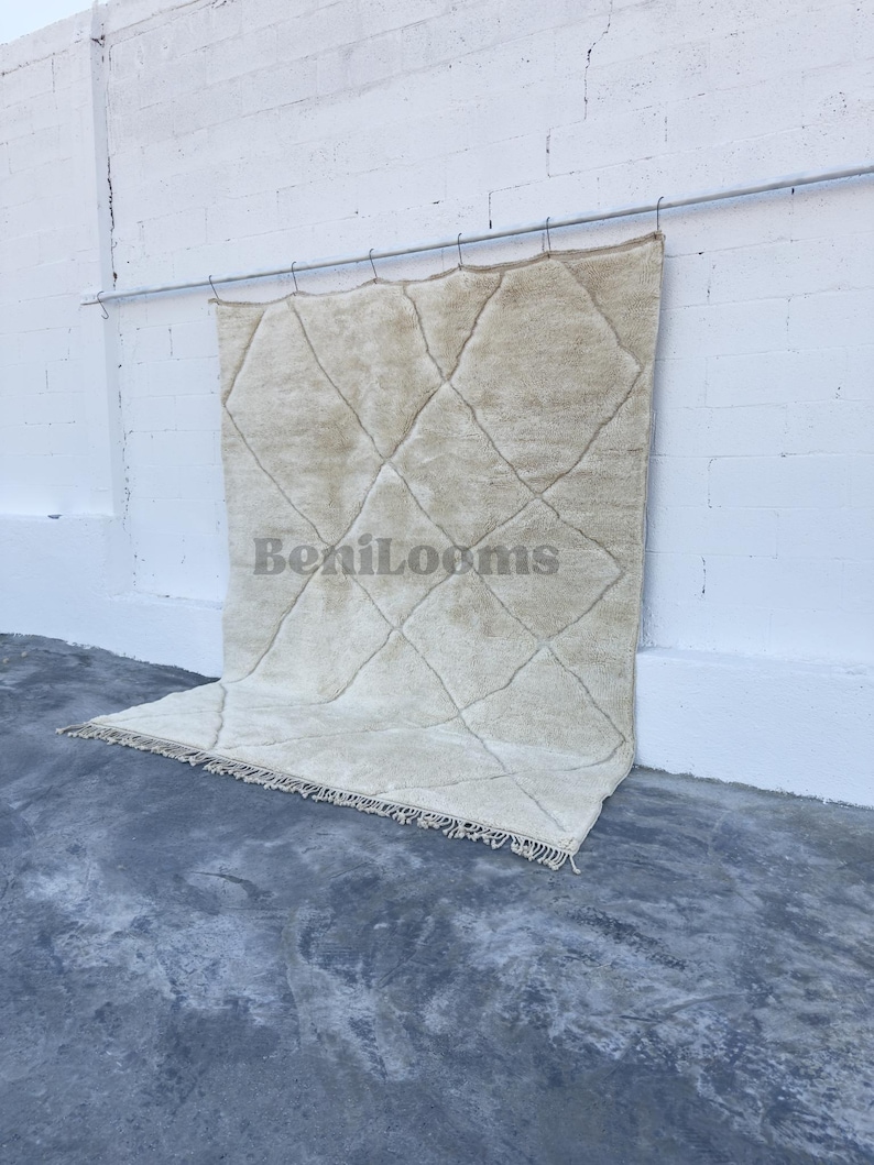 May include: A large, rectangular, cream-colored wool rug featuring a repeating diamond pattern formed by thin, dark lines. The rug has a thick, shaggy pile texture and includes a fringe along the bottom edge. The text BeniLooms is visible.