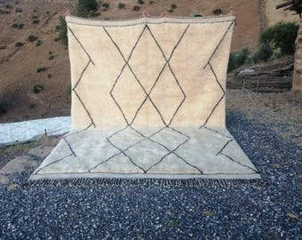 Könnte beinhalten: Ein großer, rechteckiger, cremeweißer Wollteppich mit einem sich wiederholenden schwarzen Rautenmuster auf der Oberfläche. Der Teppich hat einen dicken, hochflorigen Flor und ist an der Unterkante mit einer dunklen, geknoteten Franse versehen.