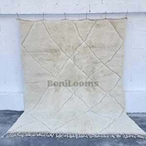 May include: A large, rectangular, off-white area rug featuring a hand-knotted, shaggy pile texture. The rug displays a prominent, dark-toned, thin diamond grid pattern across its surface and includes the text BeniLooms. It has a fringed edge.