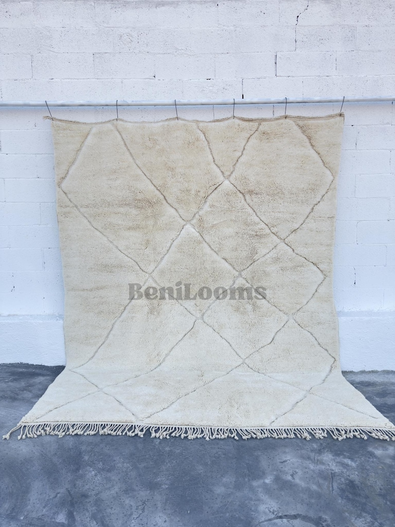 May include: A large, rectangular, off-white area rug featuring a hand-knotted, shaggy pile texture. The rug displays a prominent, dark-toned, thin diamond grid pattern across its surface and includes the text BeniLooms. It has a fringed edge.