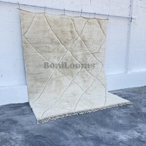 May include: A large, rectangular, cream-colored area rug featuring a dark, thin, hand-knotted diamond grid pattern across its surface. The rug has a thick, plush pile texture and includes a fringe along the bottom edge. The text BeniLooms is visible.