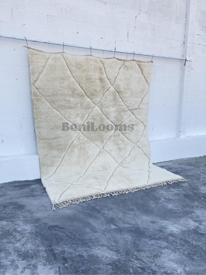 May include: A large, rectangular, cream-colored area rug featuring a dark, thin, hand-knotted diamond grid pattern across its surface. The rug has a thick, plush pile texture and includes a fringe along the bottom edge. The text BeniLooms is visible.