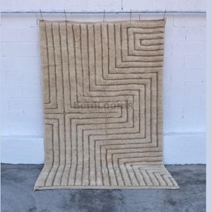 Op de afbeelding: Een rechthoekig vloerkleed in een lichtbeige kleur met een verhoogd, gestructureerd geometrisch patroon van onderling verbonden lijnen die een doolhofachtig ontwerp vormen. Het kleed heeft een dikke, hoogpolige textuur en toont BoniLooms.