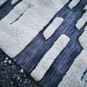 Op de afbeelding: Een wit en blauw tapijt met een geometrisch patroon. Het tapijt heeft een zachte, pluche textuur en is gemaakt van wol.