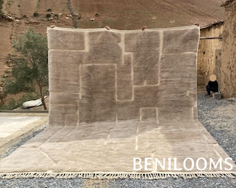 May include: A large, beige wool rug with a geometric pattern. The rug is being held up by two people, and the word "BENILOOMS" is printed on the bottom edge.