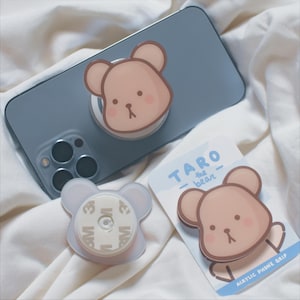 May include: A light blue phone case with a brown bear phone grip attached. The phone grip has a white adhesive backing and says "TARO - the bear - ACRYLIC PHONE GRIP".