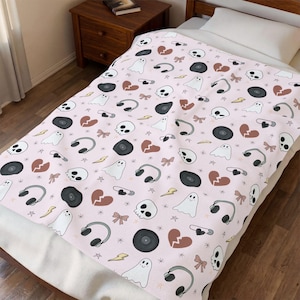 May include: A pink blanket with a repeating pattern of white ghosts, skulls, headphones, vinyl records, broken hearts, bows, safety pins, and lightning bolts. The blanket is on a bed with a wooden frame.