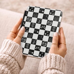 Op de afbeelding: Een zwart-wit geruit patroon hoesje met diverse grafische ontwerpen, waaronder schedels, sterren, rozen en bliksemschichten. De hoes wordt vastgehouden door een persoon die een crèmekleurige trui draagt. De hoes heeft een zwarte rand.