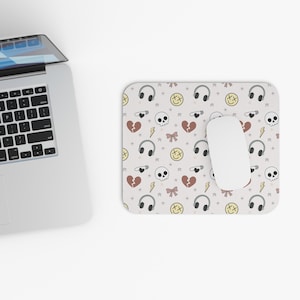 May include: A white mouse mat featuring a repeating pattern of headphones, skulls, broken hearts, bows, lightning bolts, and smiley faces. A white computer mouse sits on the mat next to a laptop.