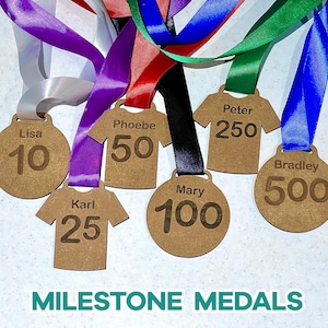 May include: A collection of milestone medals with colorful ribbons. The medals are made of wood and feature names and numbers like "Lisa 10", "Phoebe 50", and "Bradley 500". The text "MILESTONE MEDALS" is at the bottom.