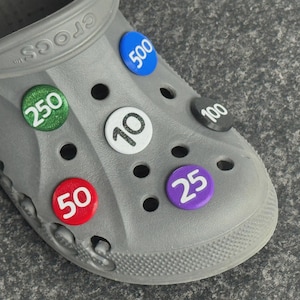 May include: A grey Crocs shoe with various colourful, circular charms. The charms display numbers: 10, 25, 50, 100, 250, and 500. The shoe has multiple holes and the word "CROCS" is visible.