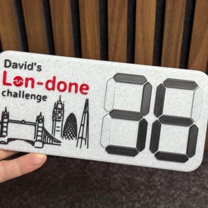 May include: A rectangular, gray sign with the text "David's Lon-done challenge" in red and black. The sign features black line drawings of London landmarks and the number "88" in a digital font.