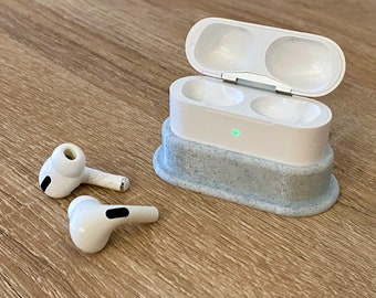 May include: White wireless earbuds and charging case. The charging case is open, revealing two earbud slots. The case sits on a speckled gray base. One earbud is outside the case, and the other is next to it.