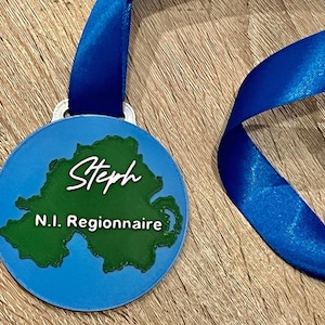 May include: A blue medal with a green map of Northern Ireland and the name "Steph" in white script. The text "N.I. Regionnaire" is below the name. A blue ribbon is attached to the medal.