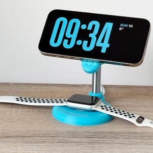 Peut inclure: Un smartphone noir affiche l'heure 09h34, monté sur un support turquoise. Une smartwatch blanche et noire avec un écran noir repose sur la base du support. La date et la température sont également affichées sur le téléphone.