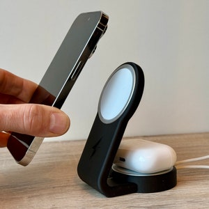May include: A black wireless charging stand with a white charging pad for a phone, a white charging pad for a watch, and a white charging pad for AirPods. The stand is on a wooden surface.